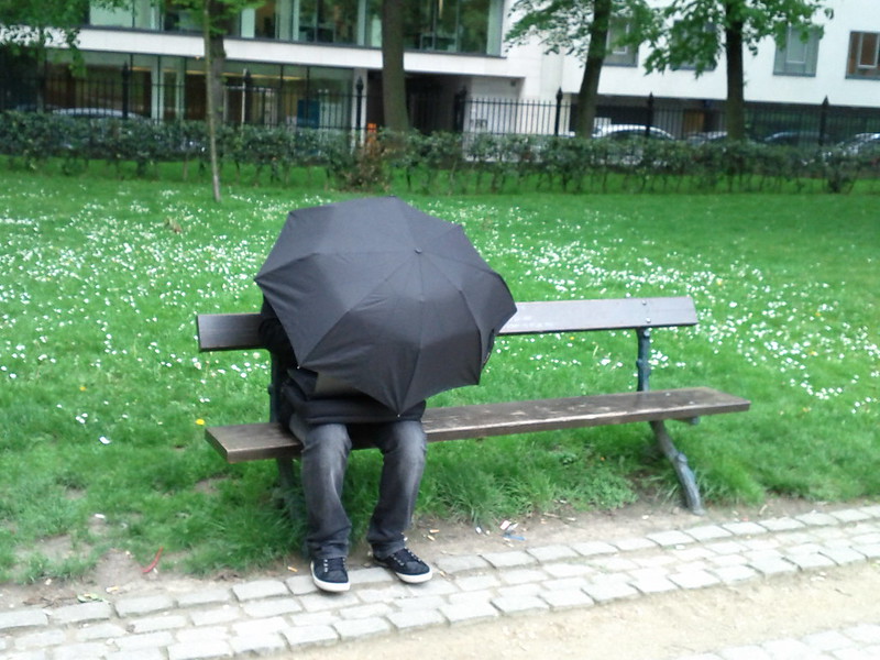 un personnage vêtu de gris assis sur un banc est presque entièrement abrité derrière un parapluie gris. le banc est sur l'herbe, au bord d'un trottoir pavé