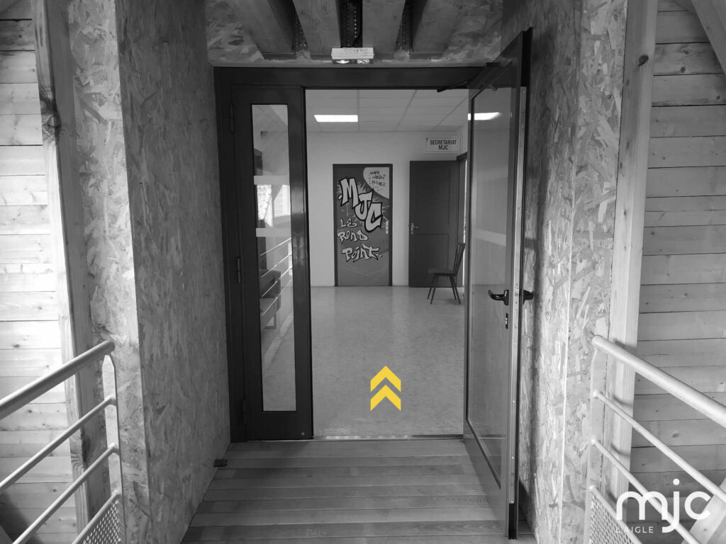 photo noir/blanc de l'entrée de la MJC, chevrons jaunes pointe vers le haut pour indiquer le sens de l'entrée. on distingue le graph sur une porte : MJC Le rond-point